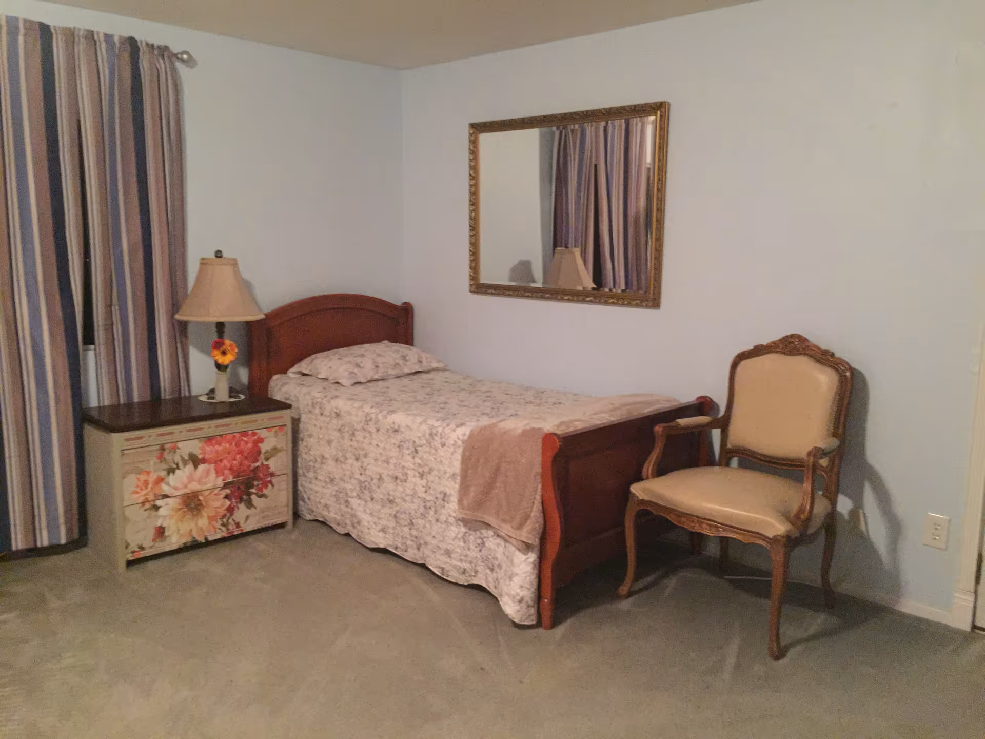 A simple bedroom with a single wooden bed covered with a floral bedspread and a beige blanket. Next to the bed is a small nightstand with a floral design, holding a lamp with a flower decoration. A large mirror with a gold frame hangs on the wall above the bed. There is a cushioned wooden chair placed beside the bed. The room has light blue walls, a window with striped curtains, and a beige carpeted floor.