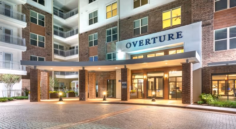 Night view of the front entrance of a multi-story senior living facility named Overture, featuring brick exterior walls, large windows, balconies, and a covered drop-off area with lights illuminating the entrance.