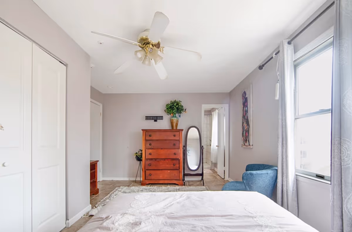 A bedroom with light gray walls, a ceiling fan, a wooden dresser with a plant on top, a full-length mirror, a blue armchair near a window with gray curtains, and a bed with a light-colored bedspread in the foreground.