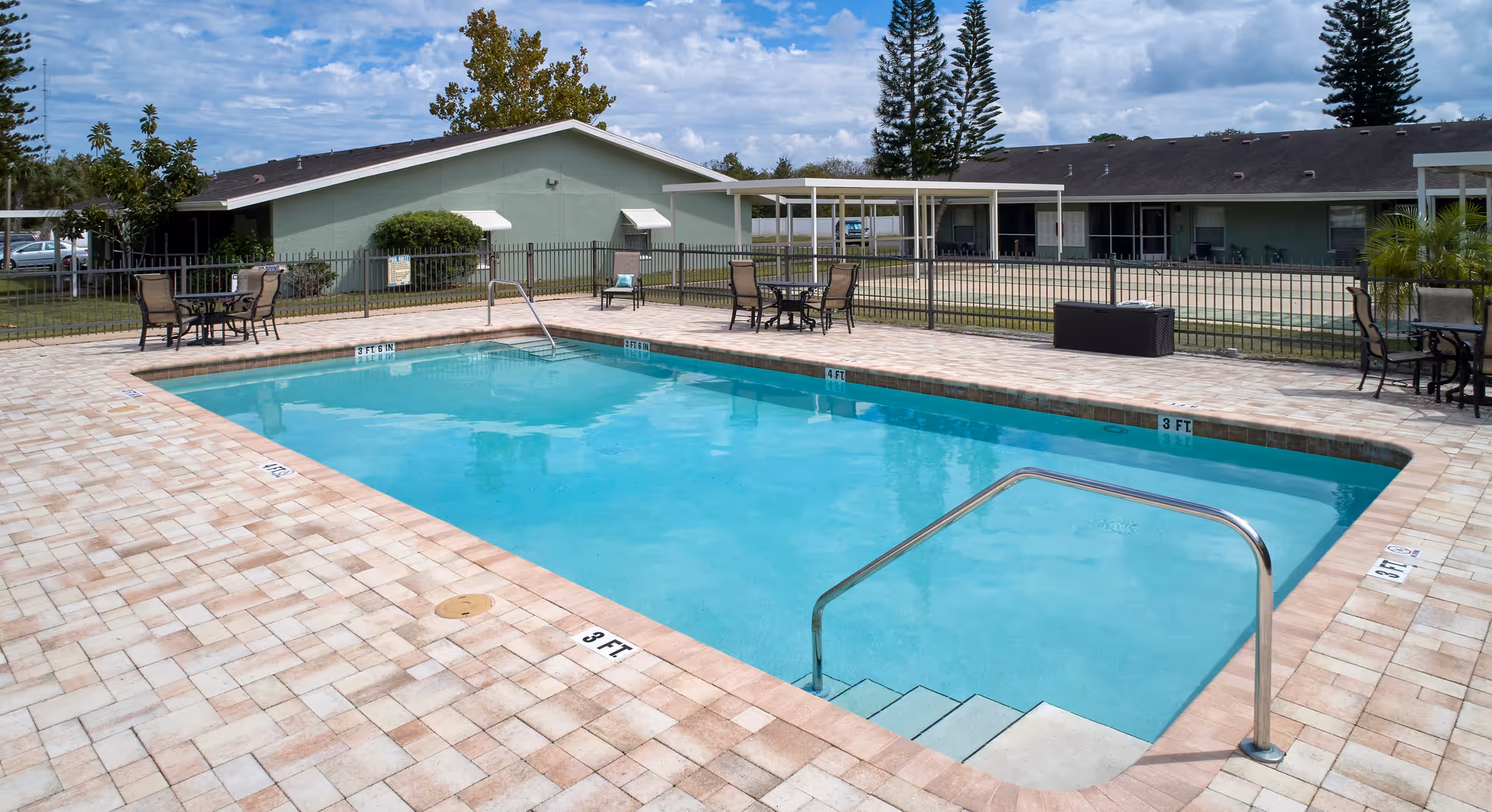 Rectangular outdoor swimming pool surrounded by a tiled patio with tables and chairs and single-story buildings in the background.