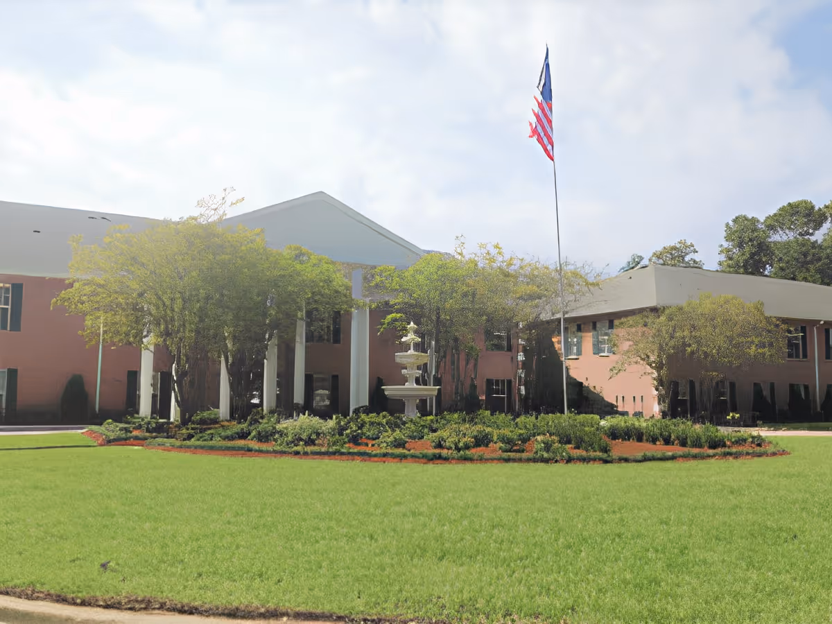 Exterior view of a senior living facility named SummerHouse Park Provence featuring a large green lawn, landscaped garden with a fountain, several trees, and an American flag on a tall flagpole in front of a two-story brick building with green window shutters.