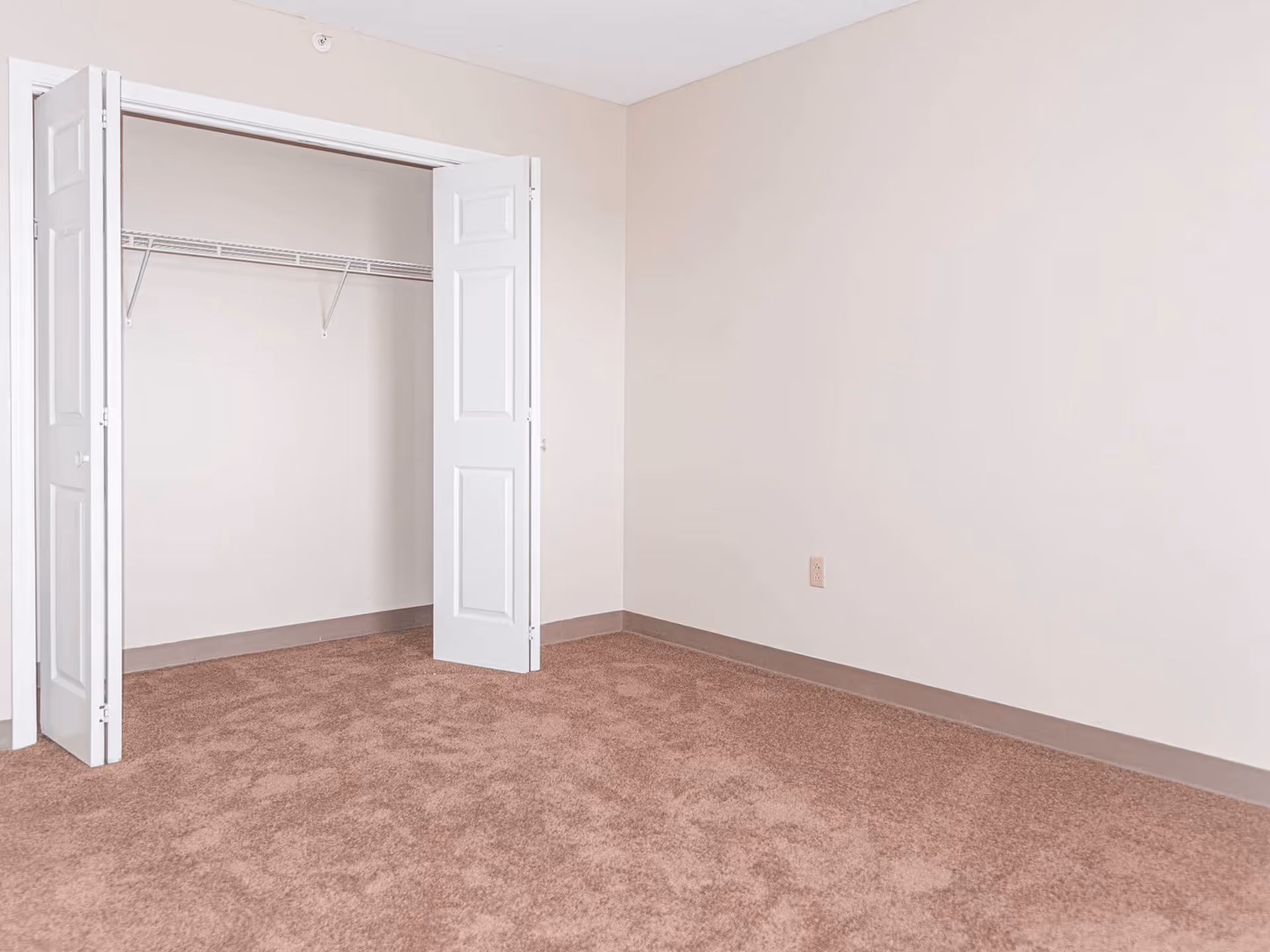 Empty room with beige walls and carpeted floor, featuring an open closet with white bi-fold doors and a wire shelf inside.
