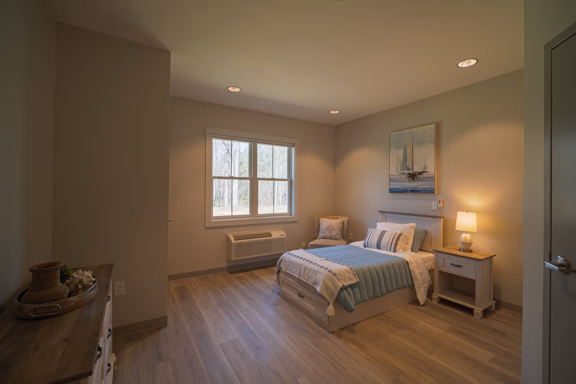A cozy bedroom with a single bed dressed in blue and white bedding, a nightstand with a lamp, a cushioned chair in the corner, a window showing trees outside, and a dresser with decorative items on top. The walls are painted light beige and there is a painting of sailboats above the bed.
