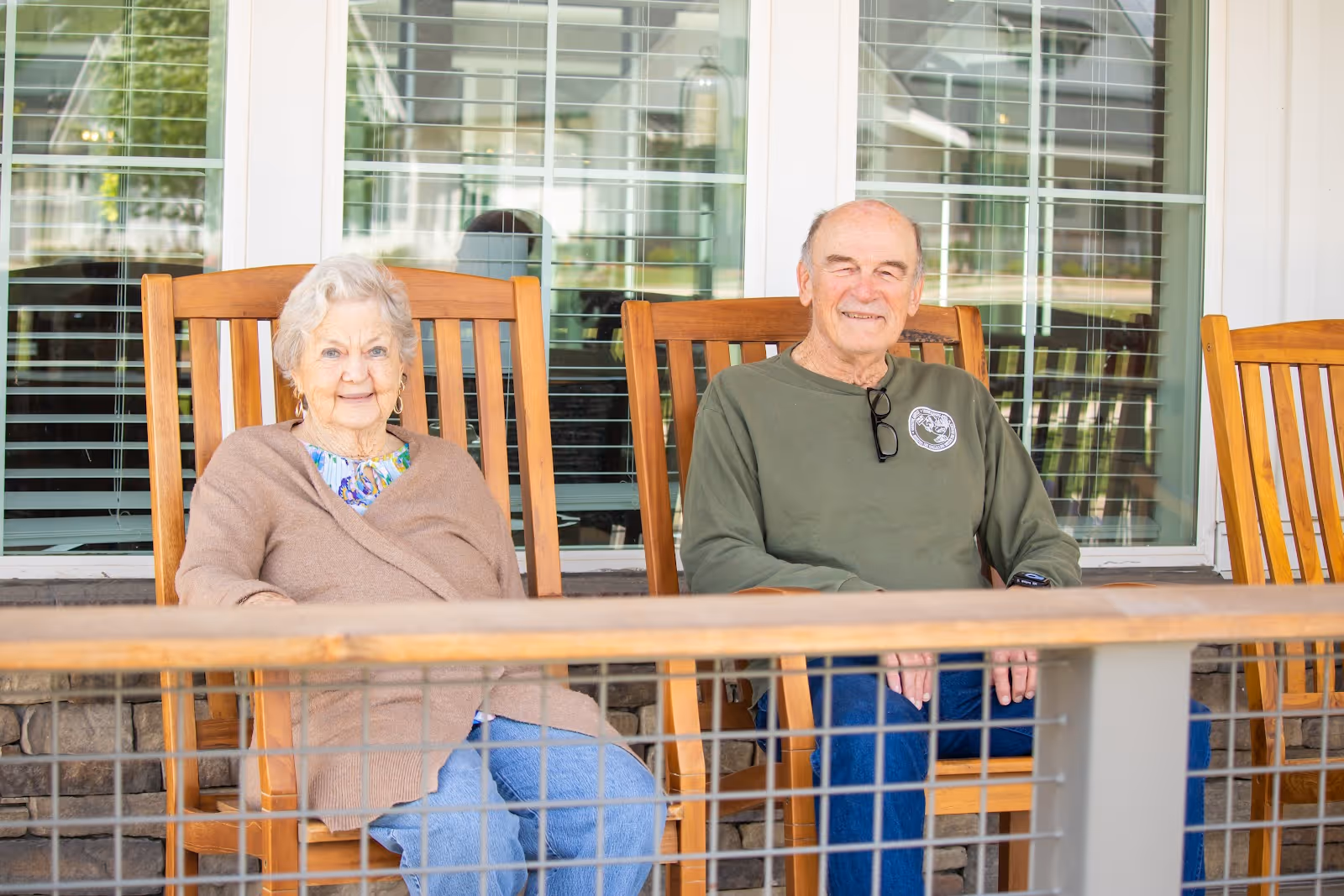 An elderly woman and an elderly man sitting on wooden rocking chairs on a porch in front of a window with white blinds. Both are smiling and appear relaxed.