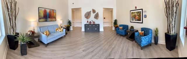 A spacious, well-lit living room area with light wood flooring and white walls. The room features a light blue sofa with decorative pillows on the left, two blue patterned armchairs with a small dark wooden table between them on the right, and a dark gray sideboard with decorative items and wall art in the center. Tall plants in black pots are placed at the corners, and colorful framed artwork hangs on the walls.