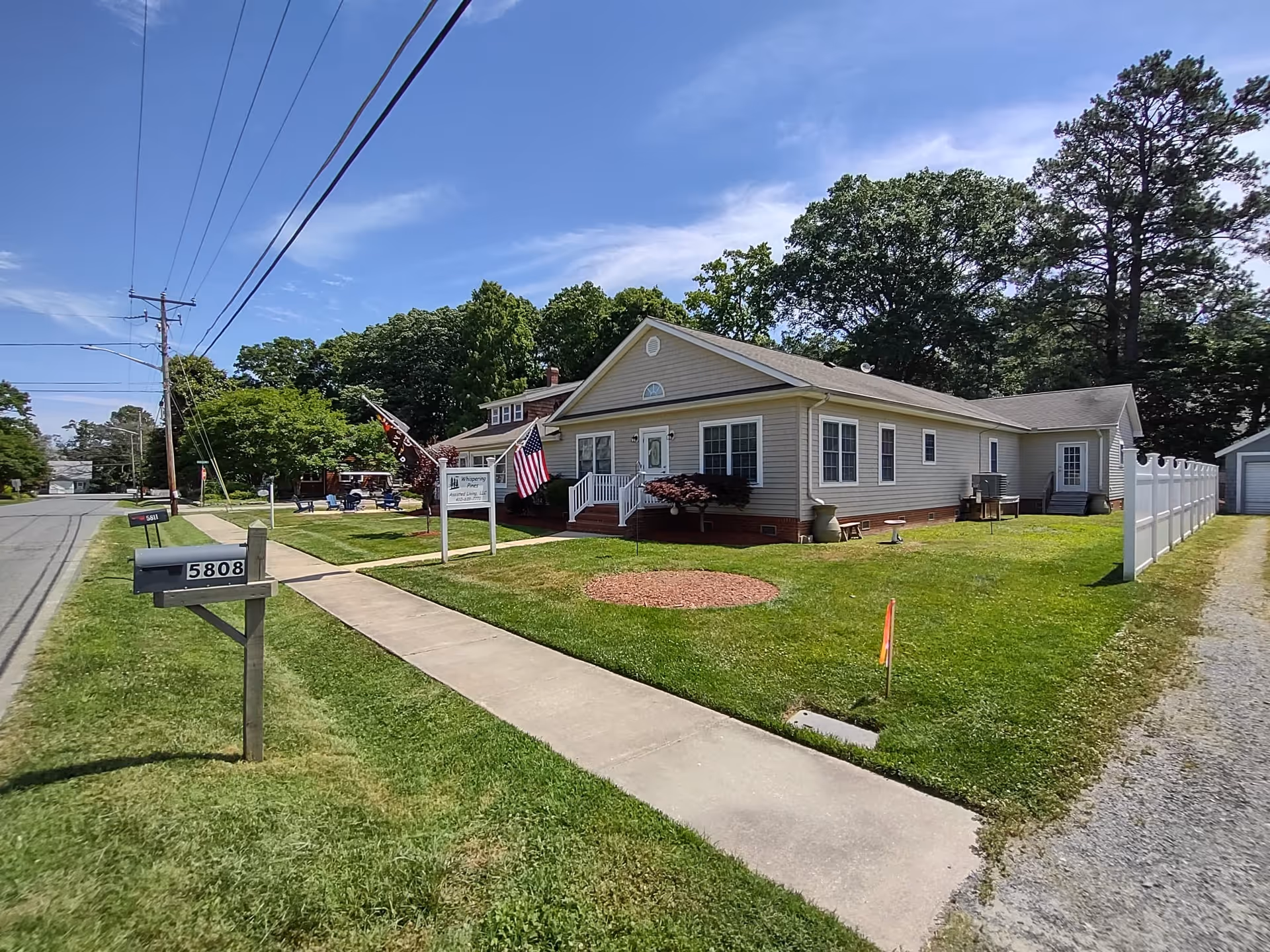 Exterior view of Whispering Pines Assisted Living Facility, a single-story beige building with white trim, a small front porch with steps, and an American flag displayed near the entrance. The building is surrounded by a well-maintained lawn, a sidewalk, a mailbox with the number 5808, and a white fence along the right side. Trees and a clear blue sky are visible in the background.