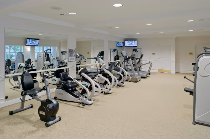 A senior living facility exercise room with several stationary exercise bikes and treadmills lined up against mirrored walls. The room has beige carpeting, white walls, and ceiling lights. Two televisions are mounted on the walls above the exercise equipment.