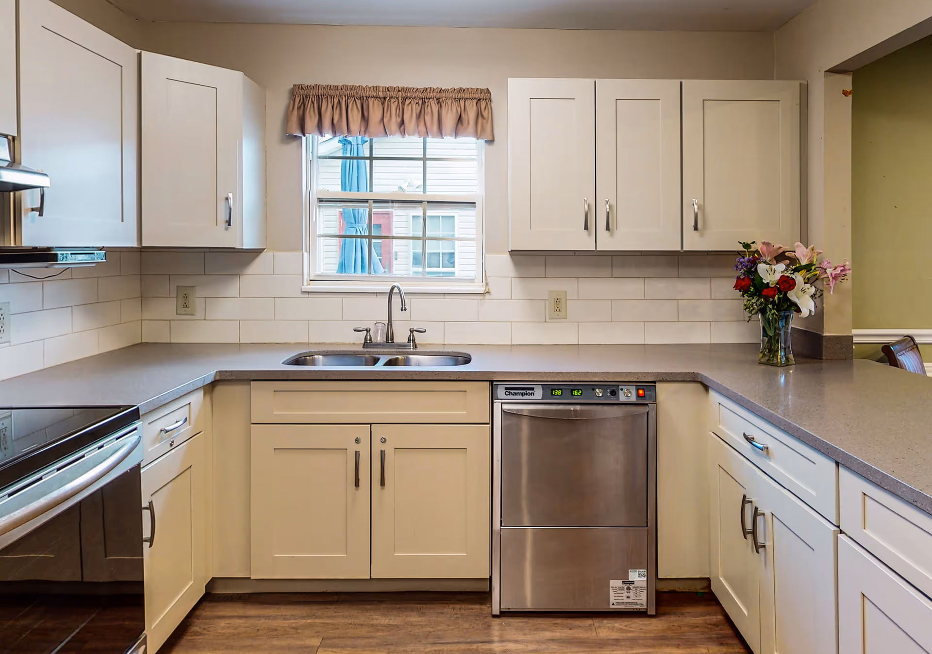 A clean and modern kitchen with white cabinets, a stainless steel dishwasher, a double sink under a window with a brown valance, and a vase of colorful flowers on the right countertop. The kitchen has a light gray countertop and white subway tile backsplash.