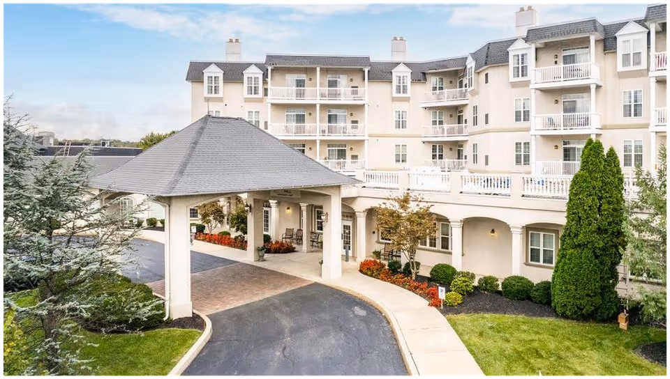 Exterior view of a multi-story senior living facility with a covered entrance driveway, landscaped greenery, and balconies on the upper floors under a partly cloudy sky.