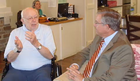 An elderly man in a wheelchair wearing a white shirt is engaged in conversation with a man in a suit and striped tie seated across from him in a room with a desk and a woman working in the background.