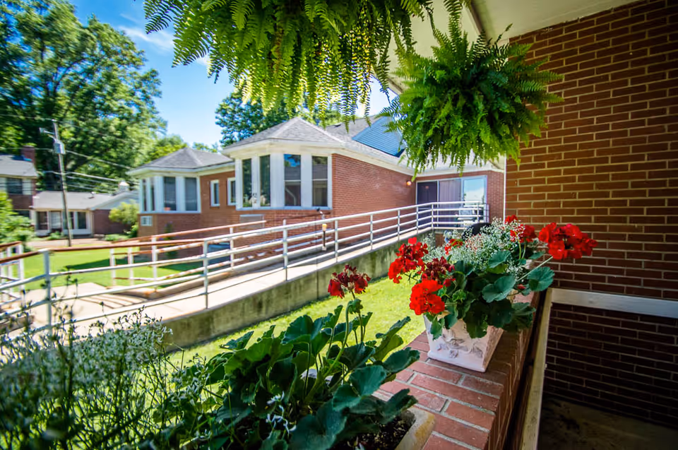 View of a brick building with a wheelchair accessible ramp, surrounded by green plants and red flowers in pots, under a bright blue sky with some trees in the background.