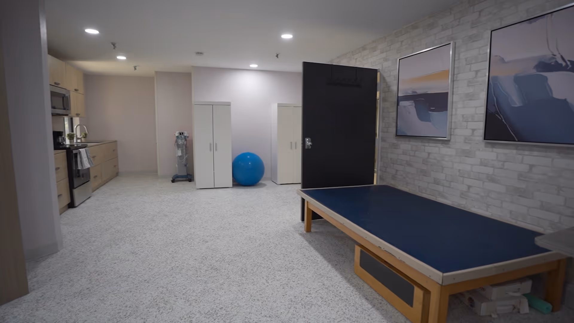 Spacious interior room with a kitchenette on the left, a therapy table on the right, storage cabinets and a blue exercise ball in the center.