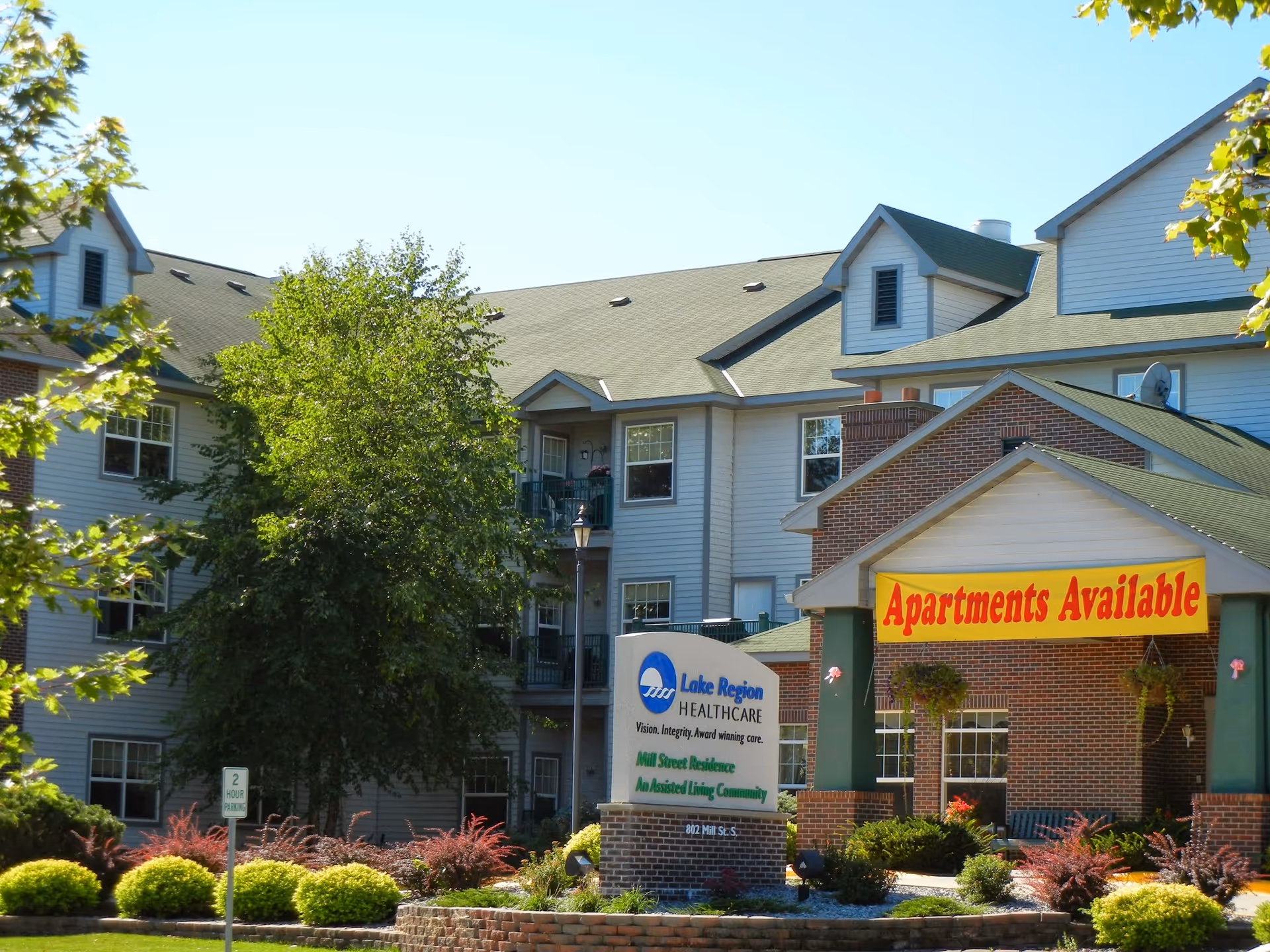 Exterior view of Mill Street Residence, a multi-story assisted living community building with a green roof and white siding. There are trees and landscaped bushes in front of the building. A sign in front reads 'Lake Region Healthcare Mill Street Residence An Assisted Living Community' and another large yellow banner on the building says 'Apartments Available'.