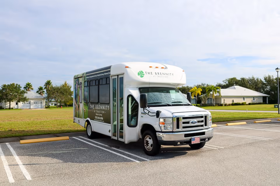 A white shuttle bus parked in an empty parking lot with grassy areas and houses in the background. The bus has signage for The Brennity at Tradition Senior Living on its side and front.
