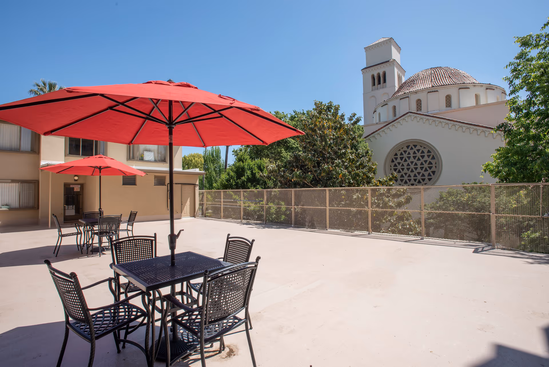Outdoor patio area at Park Place Apartments with black metal tables and chairs, each shaded by large red umbrellas. The patio is spacious with a beige concrete floor and a metal fence along the edge. In the background, there is a large building with a round window and a tower, surrounded by trees under a clear blue sky.
