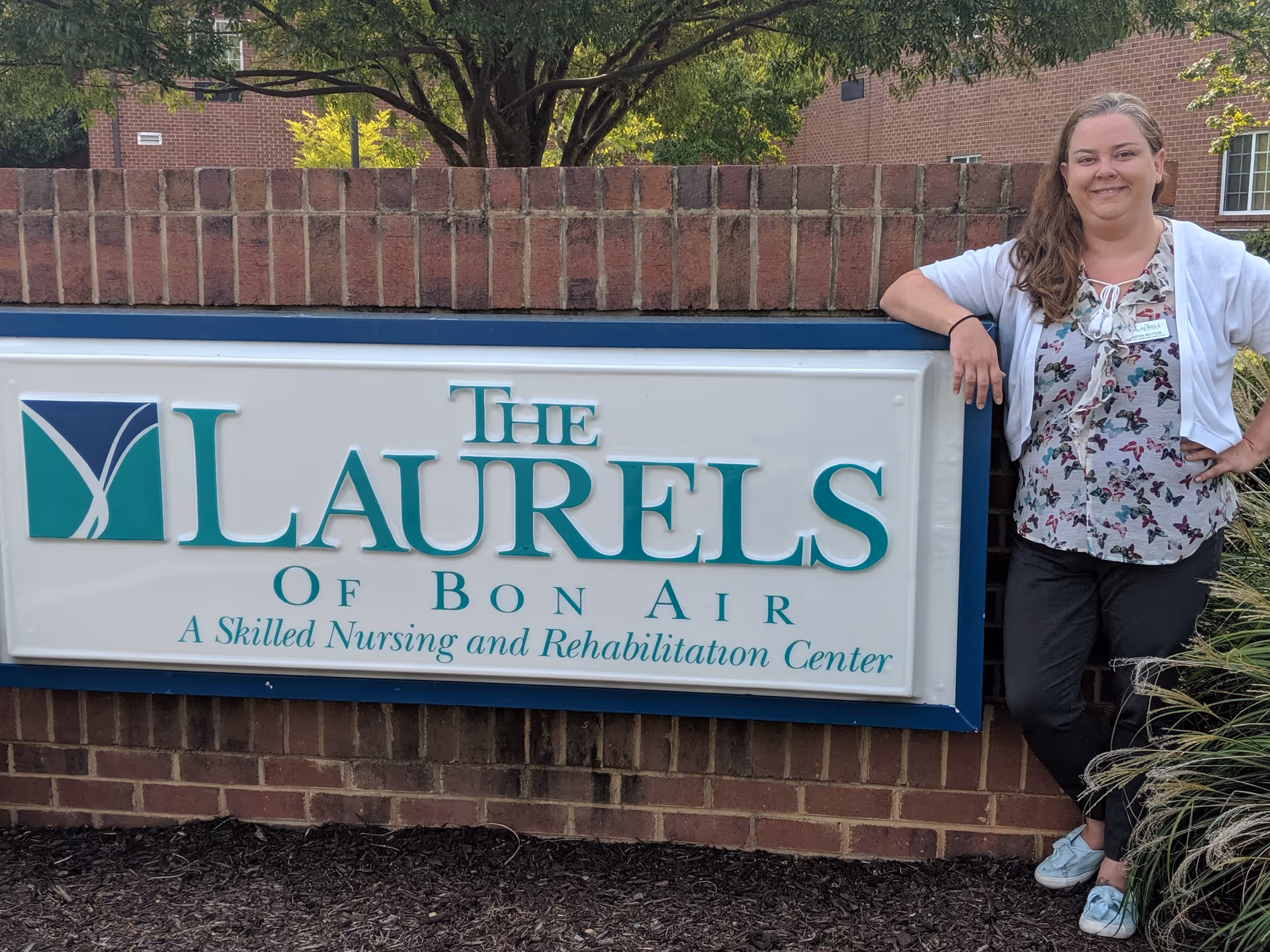 A woman stands next to a large sign for The Laurels of Bon Air, a skilled nursing and rehabilitation center. The sign is mounted on a brick wall with greenery and a building visible in the background.