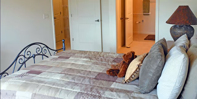 A bedroom with a neatly made bed featuring a patchwork quilt and several pillows, including a teddy bear placed on the bed. The room has a metal headboard and an adjacent lamp on a bedside table. In the background, there are two open doors leading to other rooms or closets.