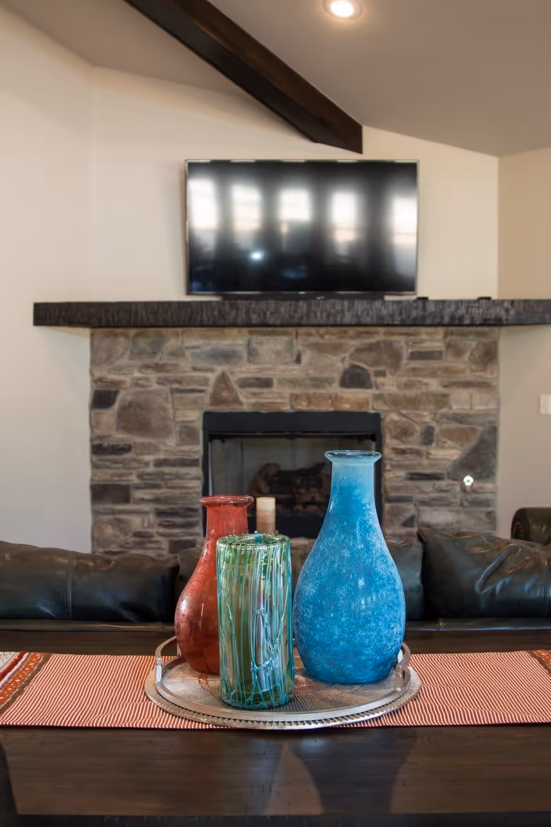 A living room interior featuring a stone fireplace with a mounted flat-screen TV above it. In front of the fireplace is a dark wooden table with a red and white striped runner, holding a silver tray with three decorative glass vases in red, green, and blue. The room has a cozy and rustic feel with exposed wooden beams on the ceiling.