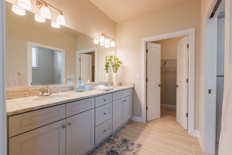 A bathroom with a long granite countertop featuring two sinks and multiple cabinets below. Above the countertop is a large mirror with two sets of three-light fixtures mounted on the wall. There are decorative items on the counter including a blue glass vase, soap dispenser, and a white vase with green plants. The floor is tiled, and there is a patterned rug in front of the sinks. A door on the right side of the image leads to a walk-in closet with wire shelving.