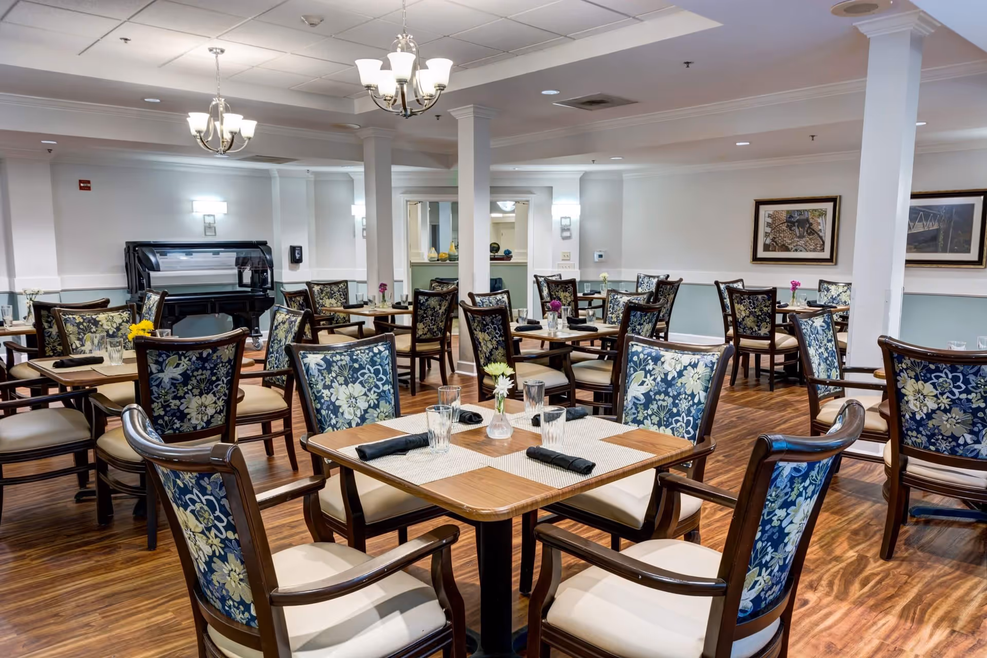 A dining room with multiple wooden tables and chairs featuring floral upholstery. Each table is set with placemats, glasses, and black napkins. The room has hardwood floors, white walls, framed artwork, and ceiling chandeliers providing lighting.