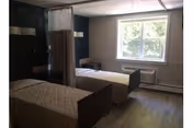 A room with two hospital-style beds separated by a privacy curtain. The beds have patterned bedspreads and are positioned near a large window letting in natural light. The room has wooden flooring and plain walls with minimal furnishings.