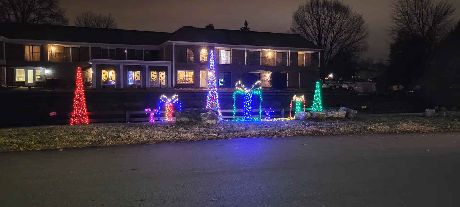 Night view of a two-story brick assisted living building front with colorful holiday lights and illuminated gift and tree shapes on the lawn.