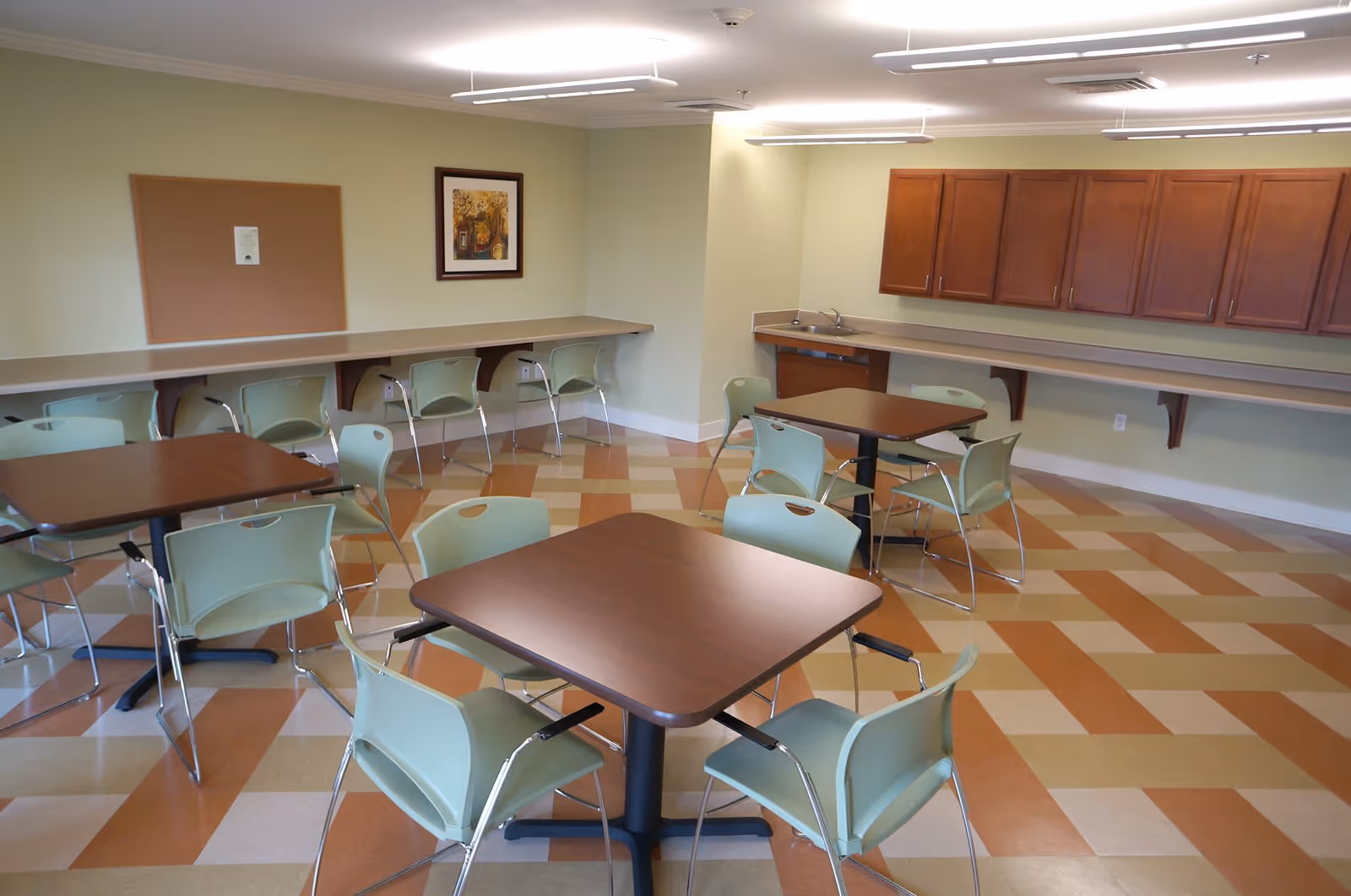 A clean and well-lit dining area with several square wooden tables surrounded by light green chairs. The room has a patterned floor with alternating beige and orange tiles. Along the walls, there are long countertops with additional chairs and wooden cabinets above a sink area. A bulletin board and framed artwork hang on the light green walls.