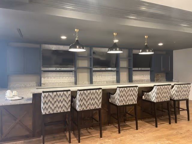 Modern kitchen area with a long marble countertop and five patterned bar stools. Above the counter are three pendant lights hanging from the ceiling. The back wall features dark cabinetry, open shelving, and three mounted flat-screen TVs. The floor is wooden.