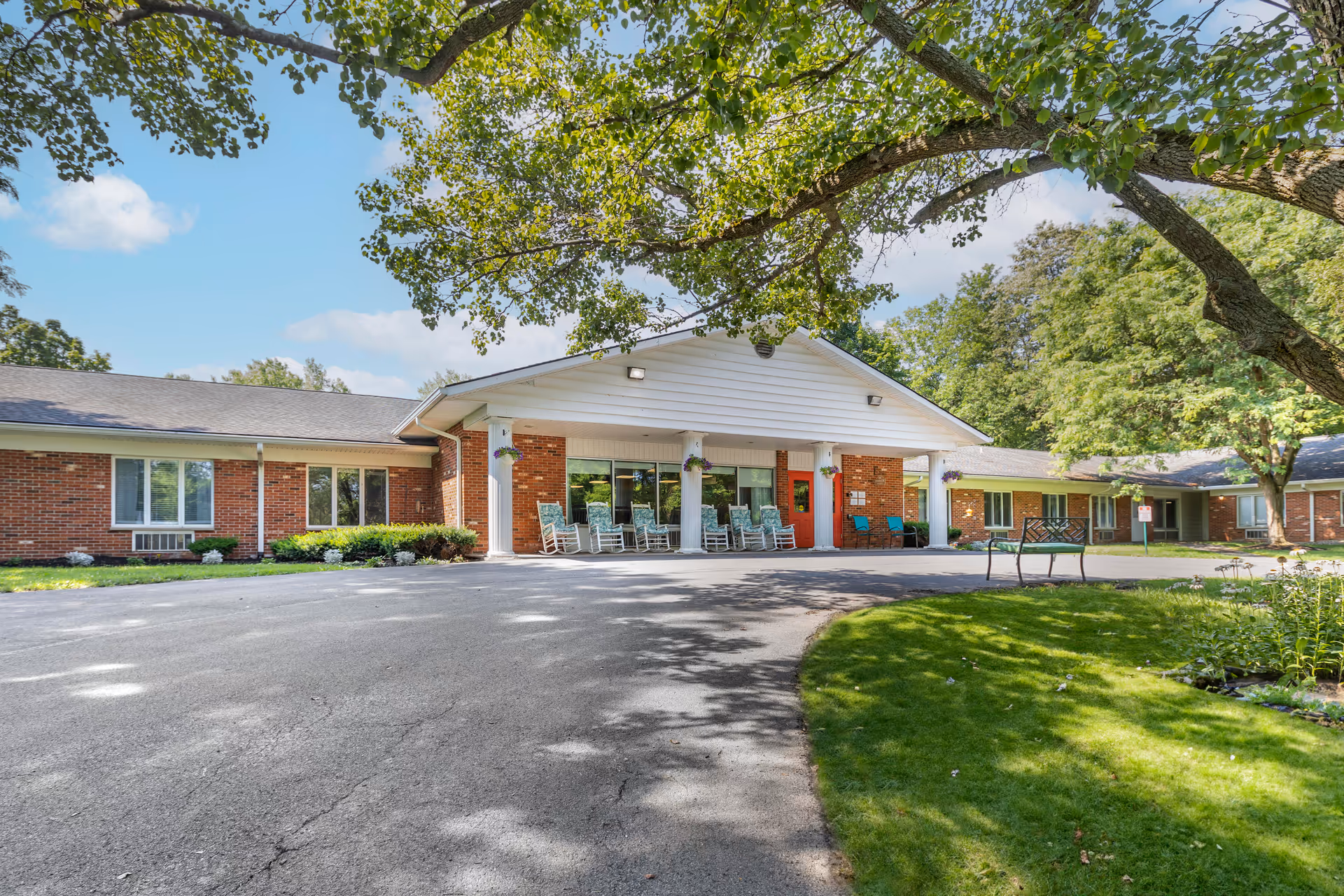 Exterior view of a single-story brick building with a covered entrance supported by white columns. Several rocking chairs are placed under the covered area. The building is surrounded by green grass, trees, and a paved driveway under a clear blue sky.