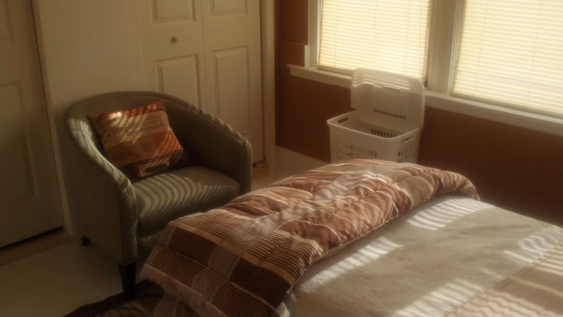 A cozy bedroom corner with a cushioned armchair featuring a patterned pillow, a bed with a brown and beige striped comforter, a white laundry basket, and windows with closed blinds letting in soft sunlight.