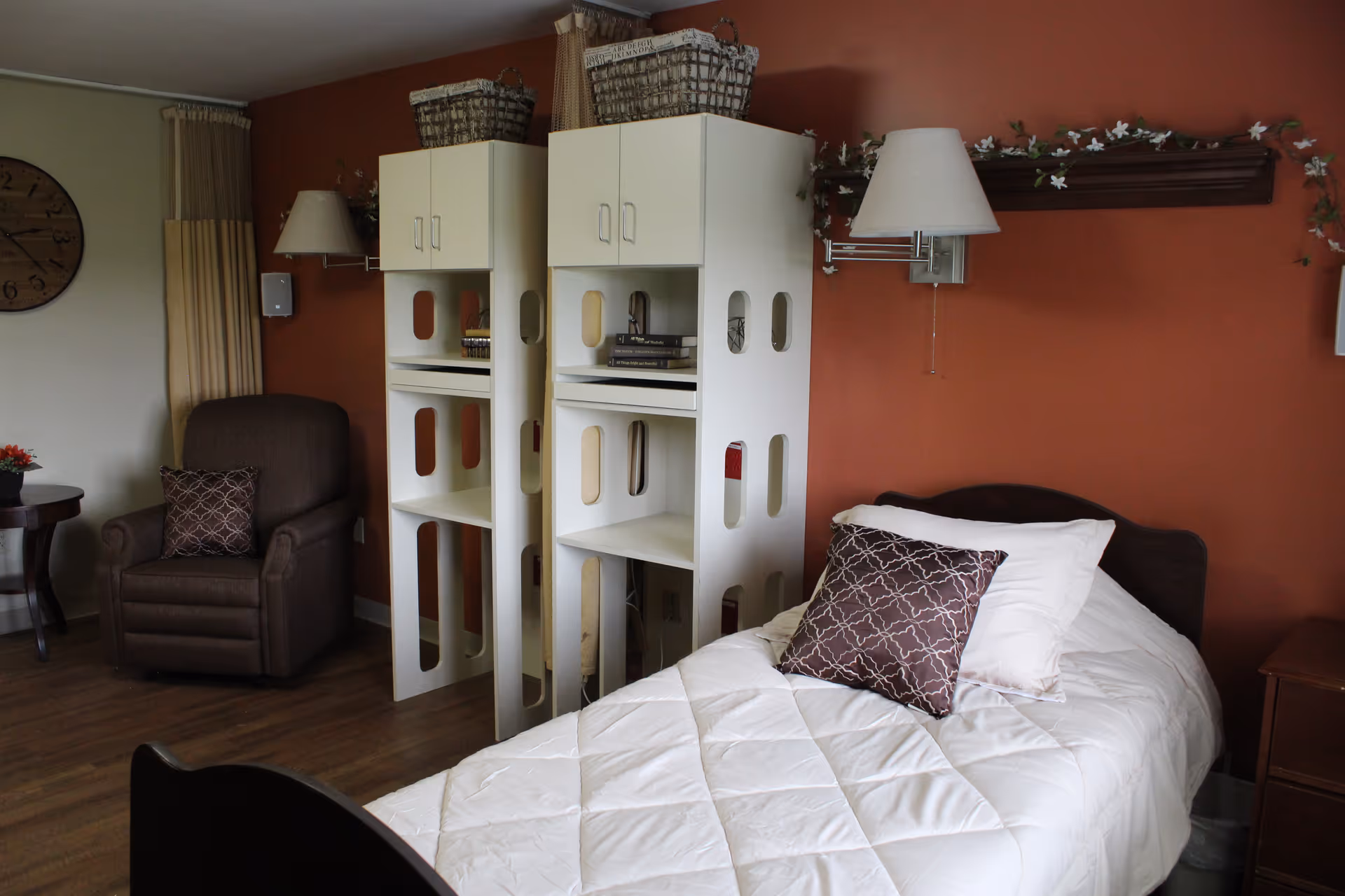 A cozy bedroom with a single bed covered in a white quilt and a decorative pillow. Behind the bed is a brown wall with a floral garland and a wall-mounted lamp. Next to the bed are two white shelving units with baskets on top and books inside. To the left, there is a brown armchair with a patterned pillow, a round side table with a small flower arrangement, and a large wall clock. The floor is wooden, and curtains hang by the window.