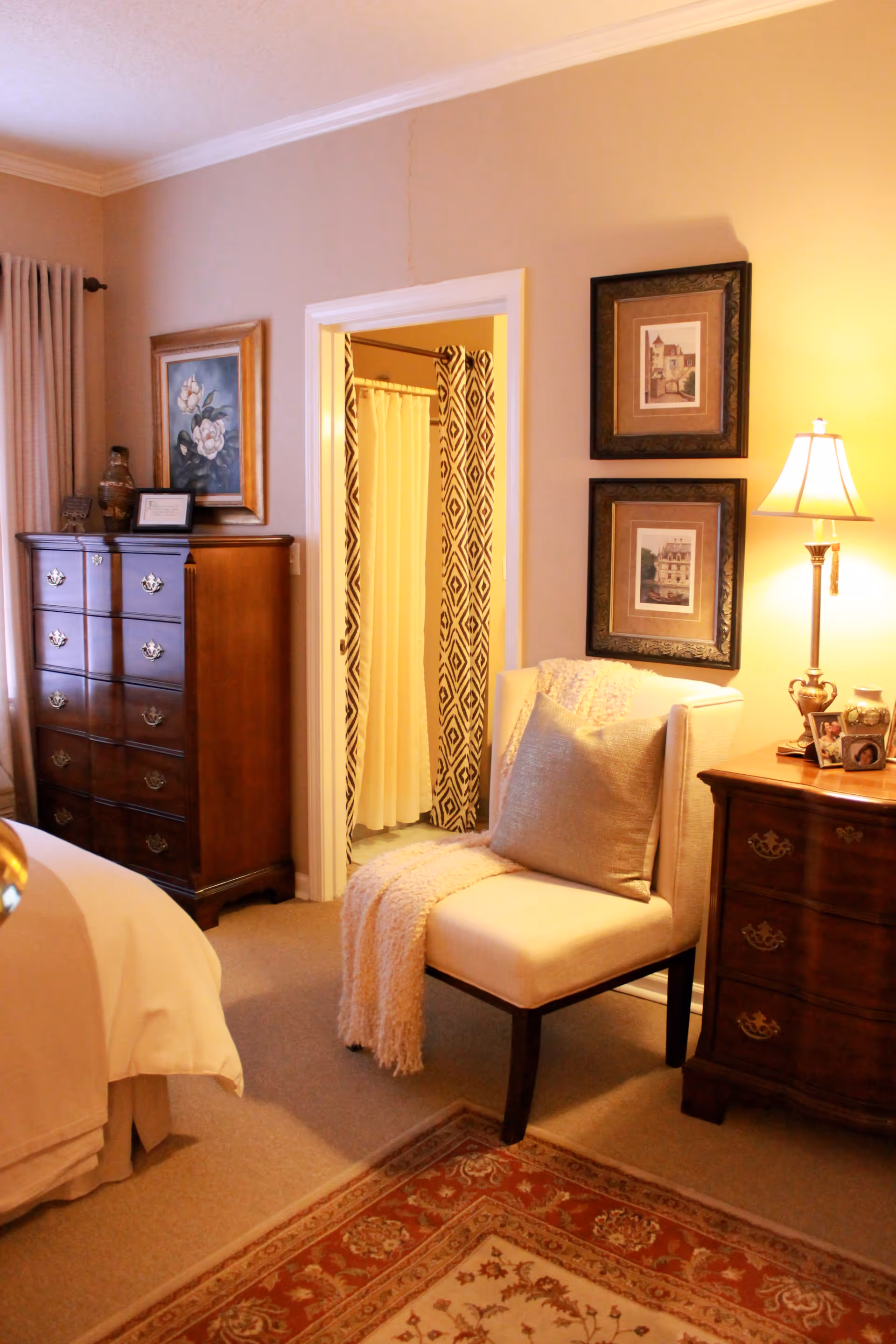 A cozy bedroom corner featuring a cushioned chair with a throw blanket and pillow, a wooden nightstand with a lamp and decorative items, two framed pictures on the wall, a wooden dresser with a framed picture and decorative vase, and an open doorway leading to a bathroom with a patterned shower curtain.
