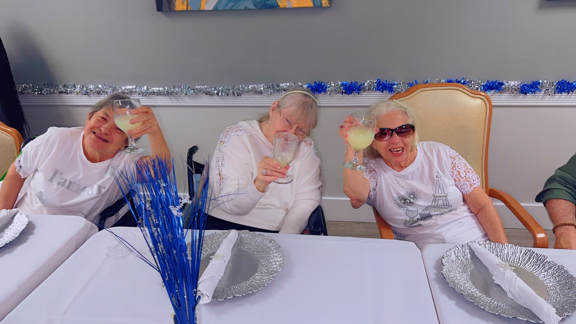 Three elderly women sit at a decorated table in a dining room, smiling and raising glasses in a toast.