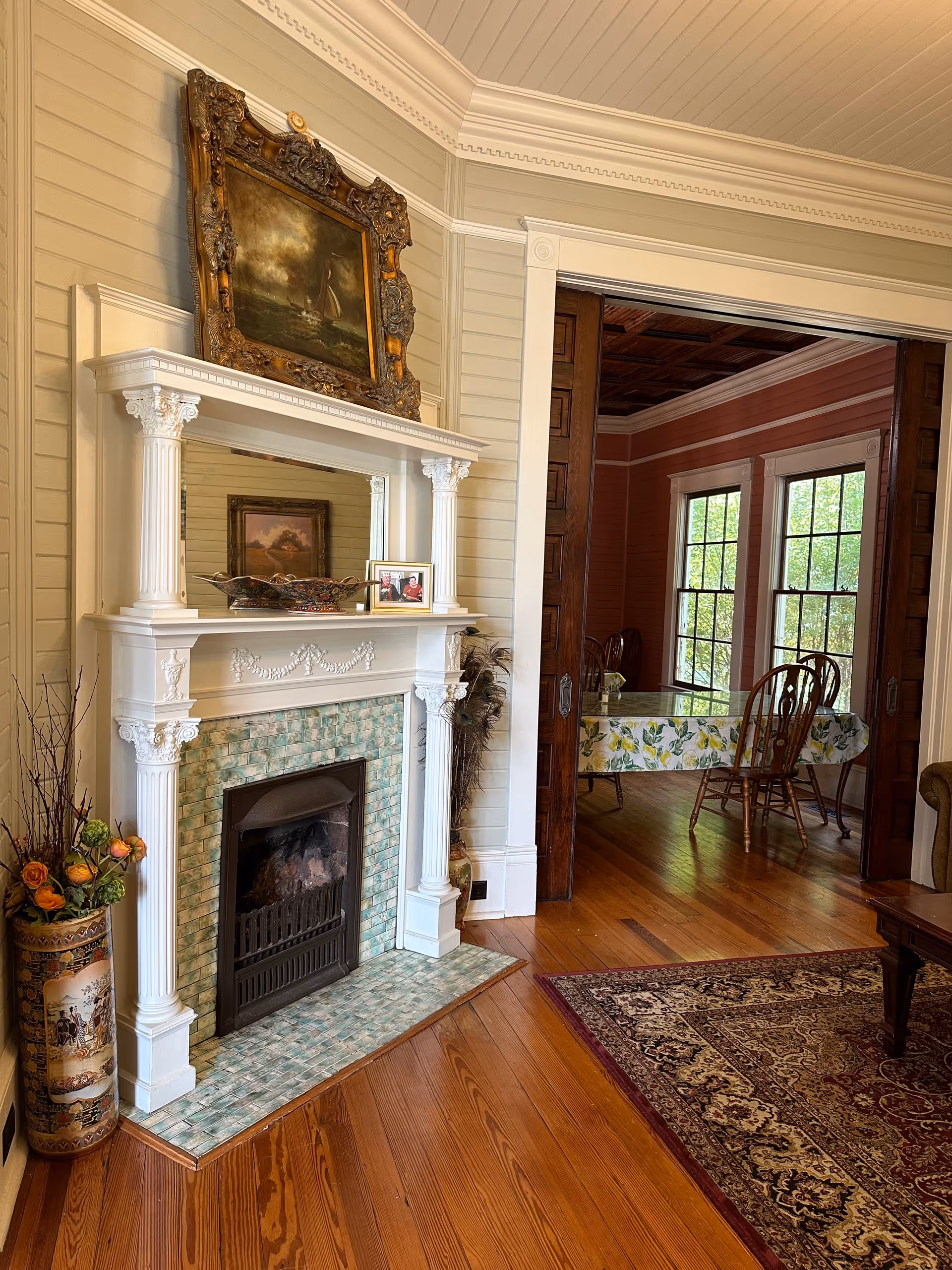A cozy interior scene featuring a white ornate fireplace with green tiles and a large framed painting above it. On the mantel are decorative items including a framed photo and a bowl. To the left of the fireplace is a tall decorative vase with flowers. The room has wooden floors and cream-colored walls with detailed molding. Through an open doorway with wooden sliding doors, a dining area with a table covered in a floral tablecloth and wooden chairs is visible, with large windows letting in natural light.