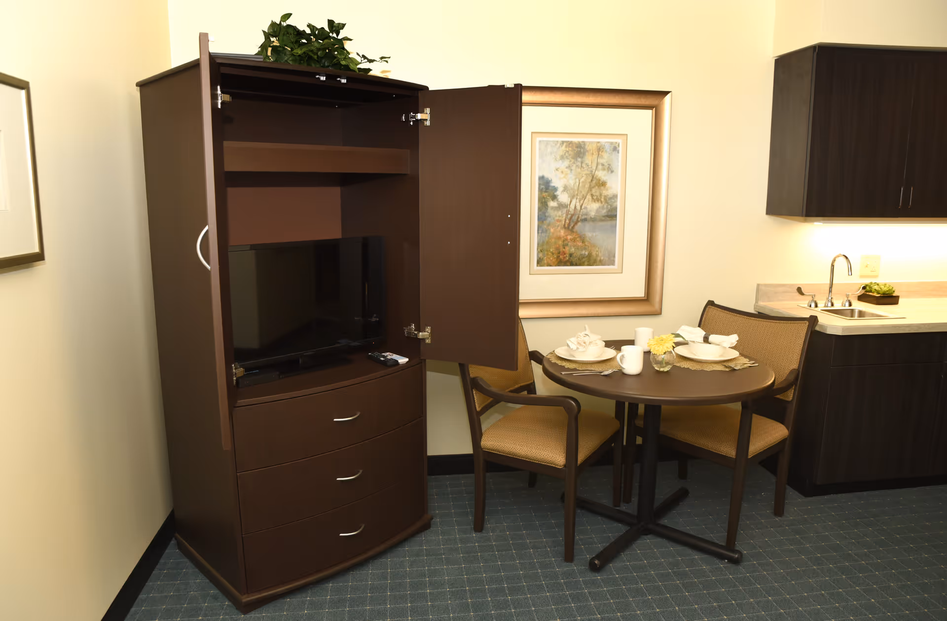 A small dining area in a senior living facility with a round table set for two, two chairs, a dark wood cabinet with open doors revealing a television inside, and a kitchenette with a sink and cabinets. A framed landscape painting hangs on the wall behind the table.