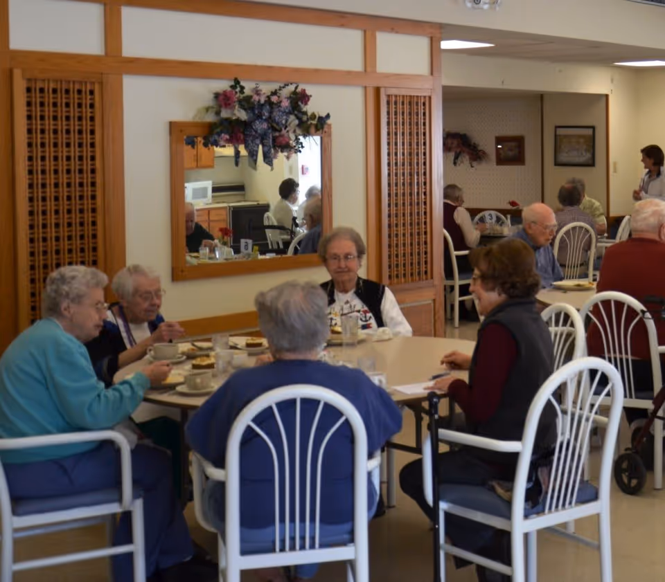 A group of elderly people sitting around tables in a dining area, eating and conversing. The room has white walls, wooden accents, and a mirror with floral decorations above it. Several other elderly individuals are visible in the background, along with a staff member standing near the wall.