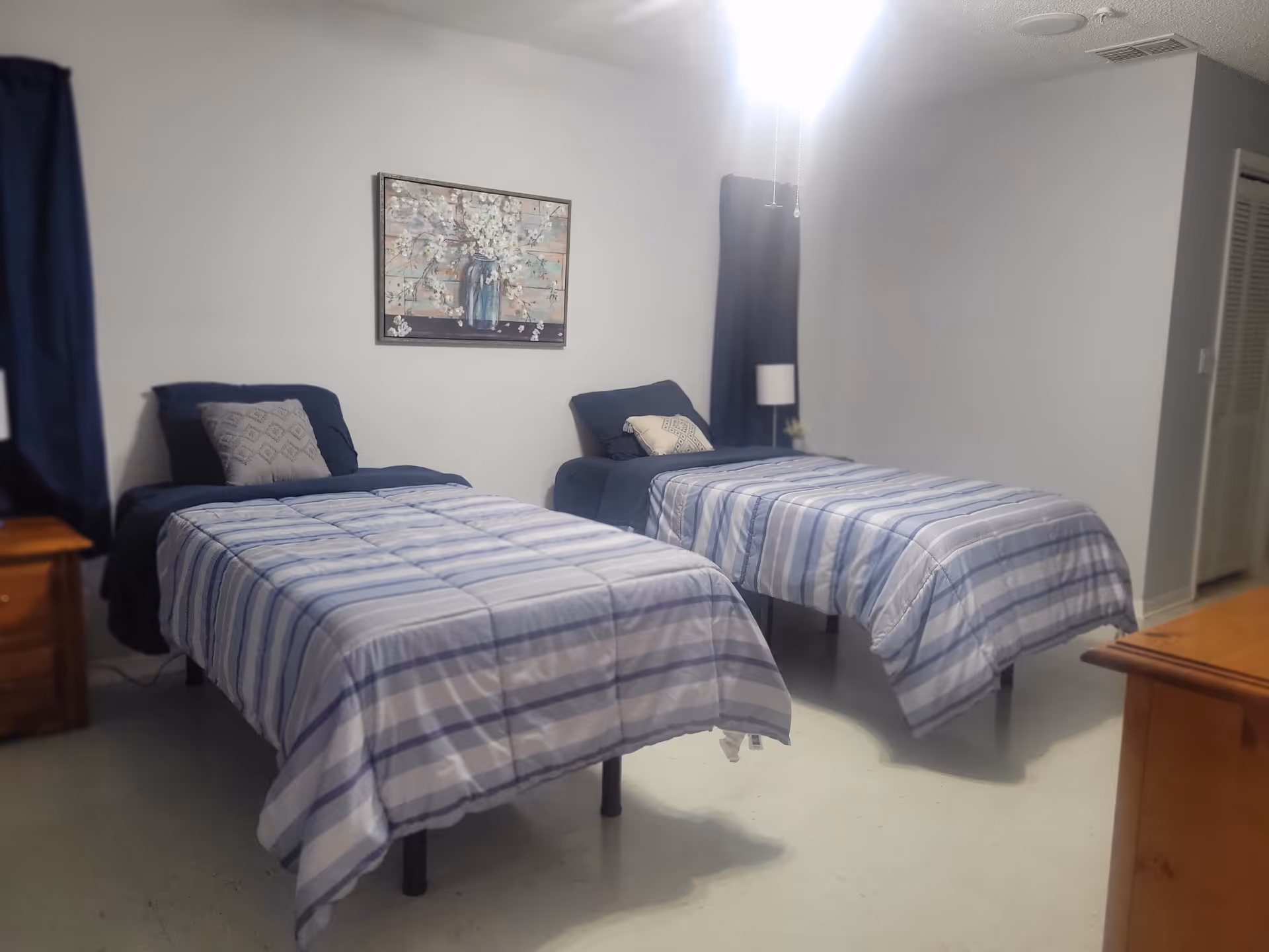 A bedroom with two twin beds side by side, each with blue and white striped bedding and pillows. There is a wooden nightstand with a lamp between the beds, a framed floral painting on the wall above the beds, and a window with dark curtains on the left side. The room has light-colored walls and flooring.