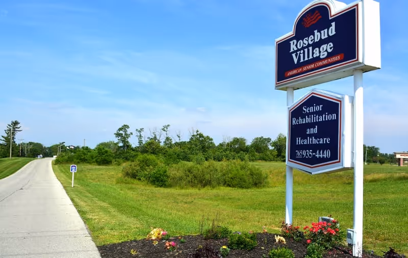 Entrance sign for Rosebud Village reading 'Senior Rehabilitation and Healthcare' beside a road and grassy field under a blue sky.