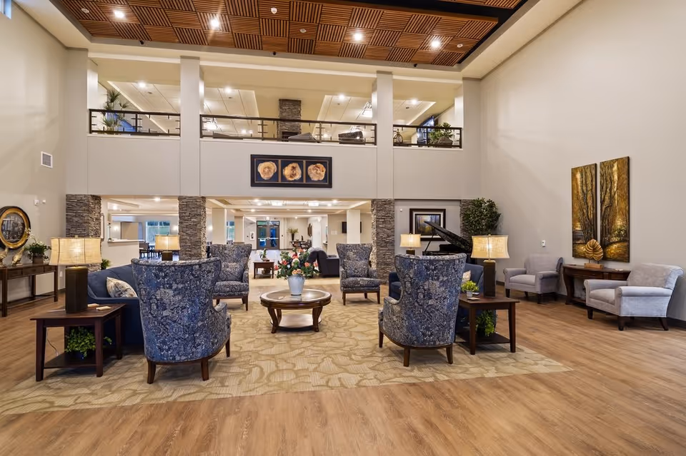 Spacious senior living facility common area with high ceilings, featuring a seating arrangement of blue patterned armchairs around a round wooden coffee table with a flower vase. The room has wooden flooring, beige walls, decorative paintings, table lamps, and a grand piano in the background. There is a second-floor balcony overlooking the area.