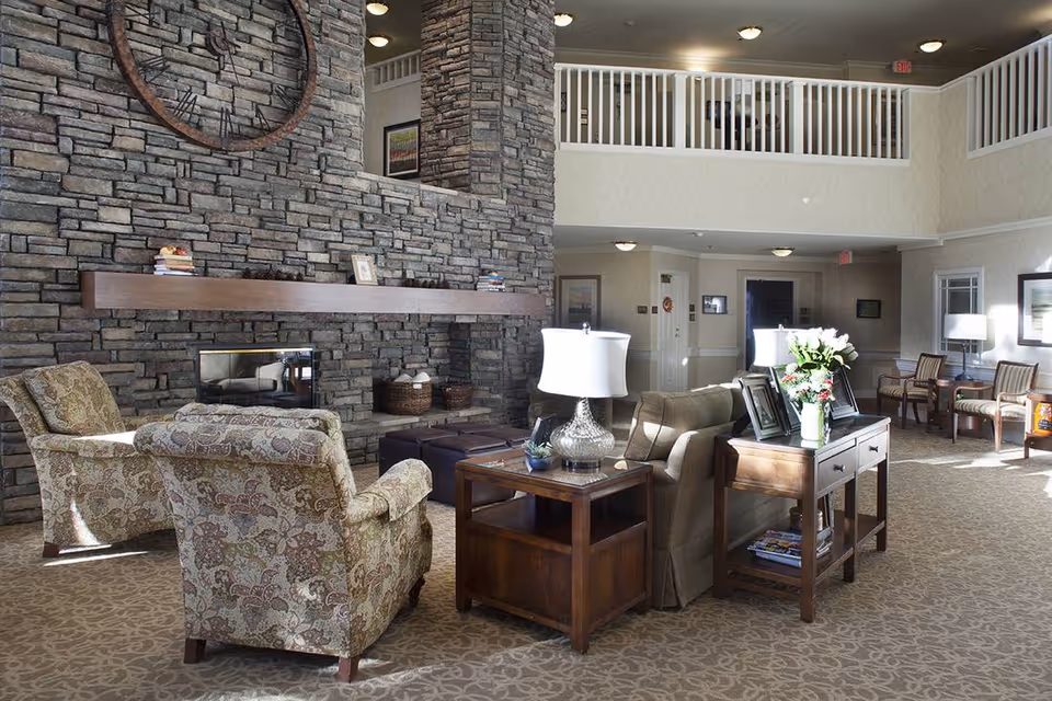 A spacious and well-lit living room area with a high ceiling and a stone fireplace. The room features comfortable upholstered armchairs and a sofa arranged around wooden side tables with lamps and decorative items. There is a large circular mirror above the fireplace and a balcony railing visible on the upper level. The carpet has a subtle floral pattern, and the walls are painted in light neutral tones.