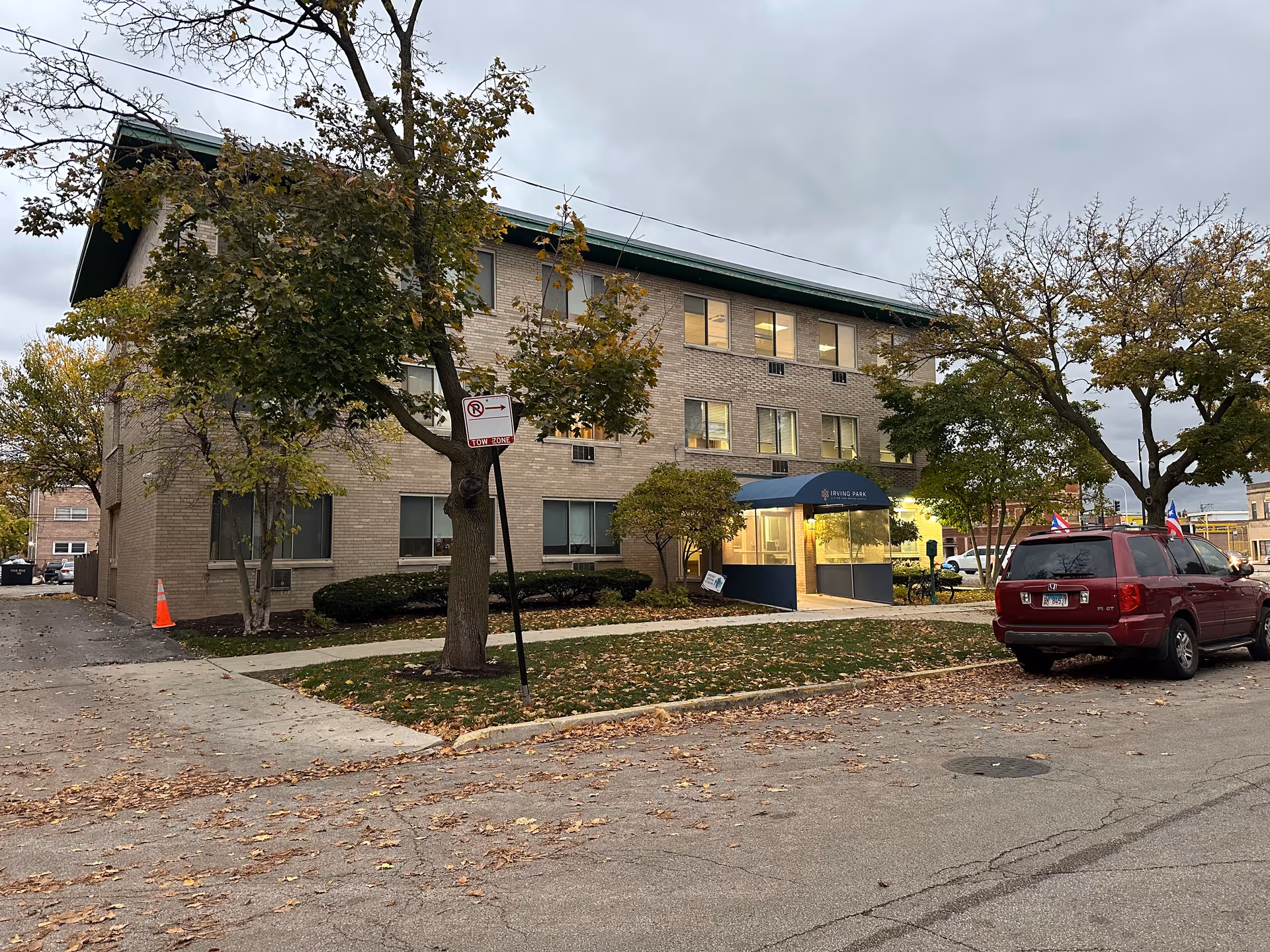 Exterior view of a three-story brick building with a blue awning entrance labeled Irving Park Living and Rehab Center. There are trees with autumn leaves around the building, a red SUV parked on the street, and a no parking sign near the sidewalk.