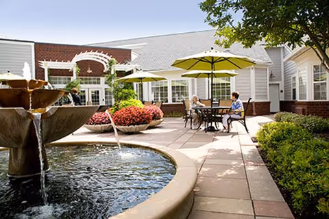 Outdoor courtyard area at Parc Provence featuring a water fountain in the foreground, patio tables with umbrellas, and two elderly women sitting and conversing. The courtyard is surrounded by a single-story building with large windows and greenery.