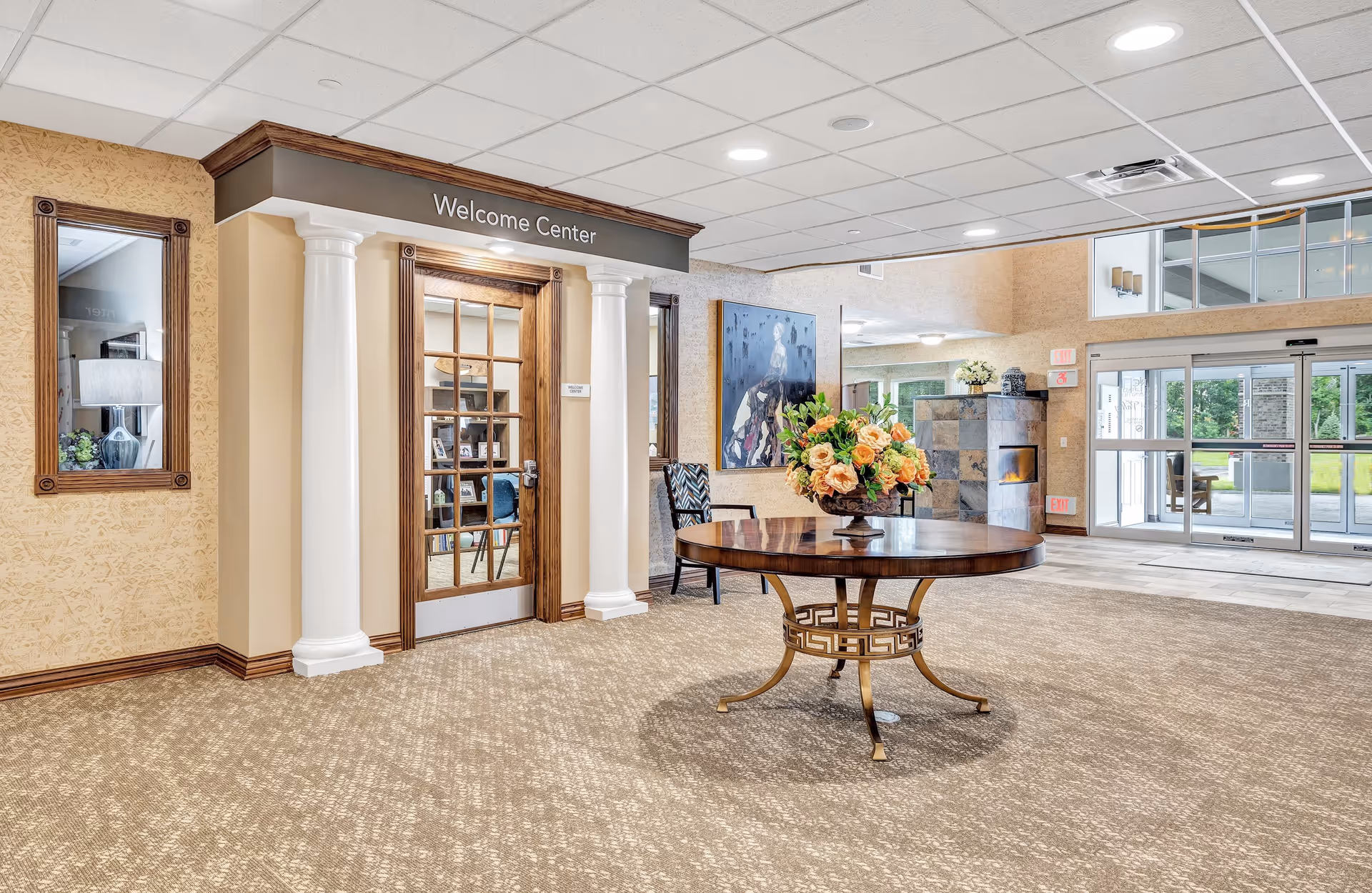 Interior view of a retirement community's entrance area featuring a round wooden table with a floral arrangement in the center. To the left is a door labeled 'Welcome Center' framed by two white columns. The area has beige carpet, light-colored walls, and a large windowed entrance door on the right side allowing natural light in. A painting and a chair are visible near the Welcome Center door.