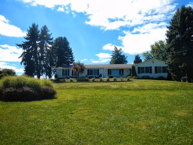 Single-story white house on a grassy hill with trees under a bright blue sky.