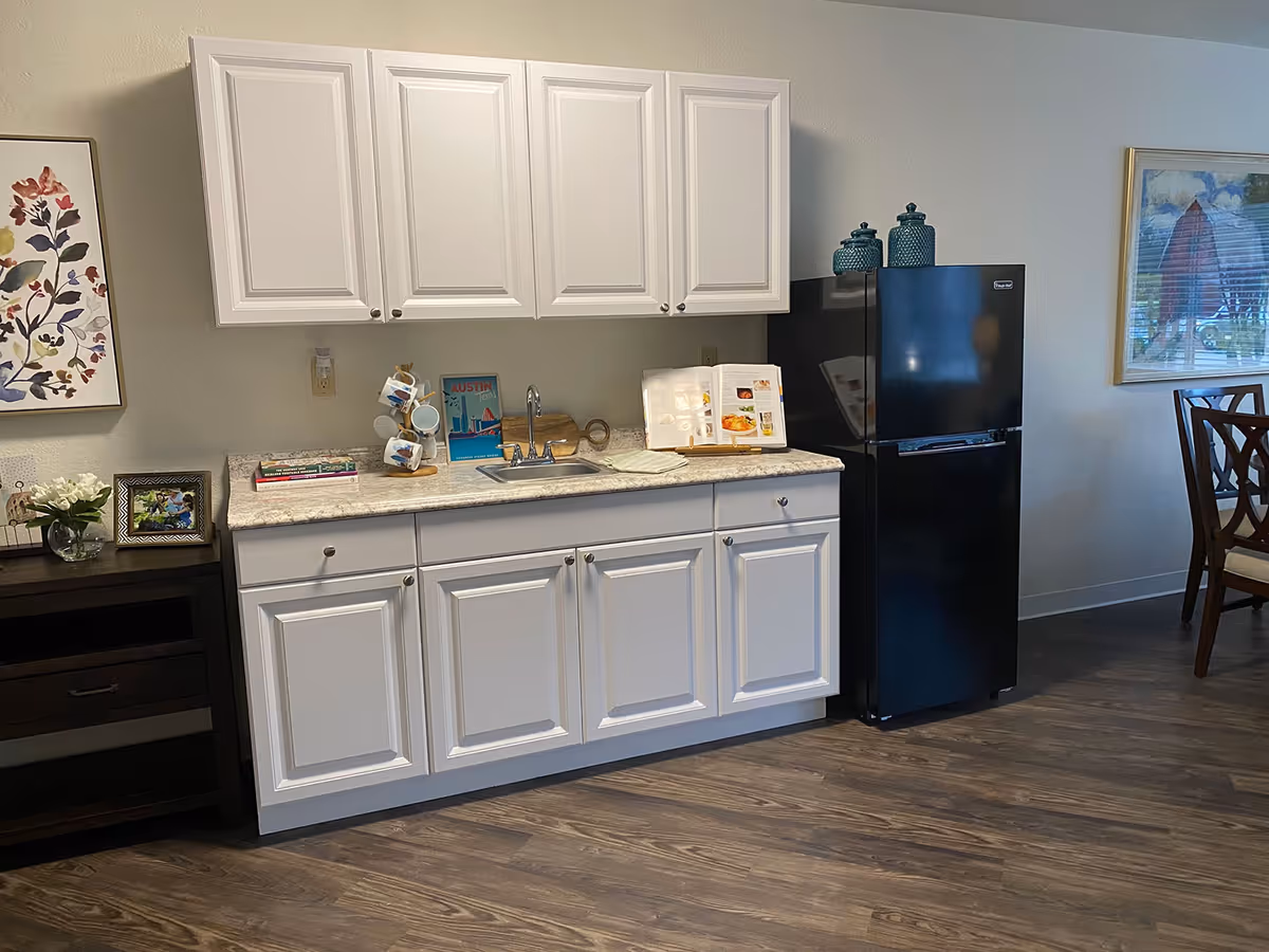 A small kitchen area with white cabinets, a countertop with a sink, a black refrigerator, and decorative items including a mug tree, books, and framed pictures. There is a wooden floor and part of a dining table with chairs is visible on the right side.