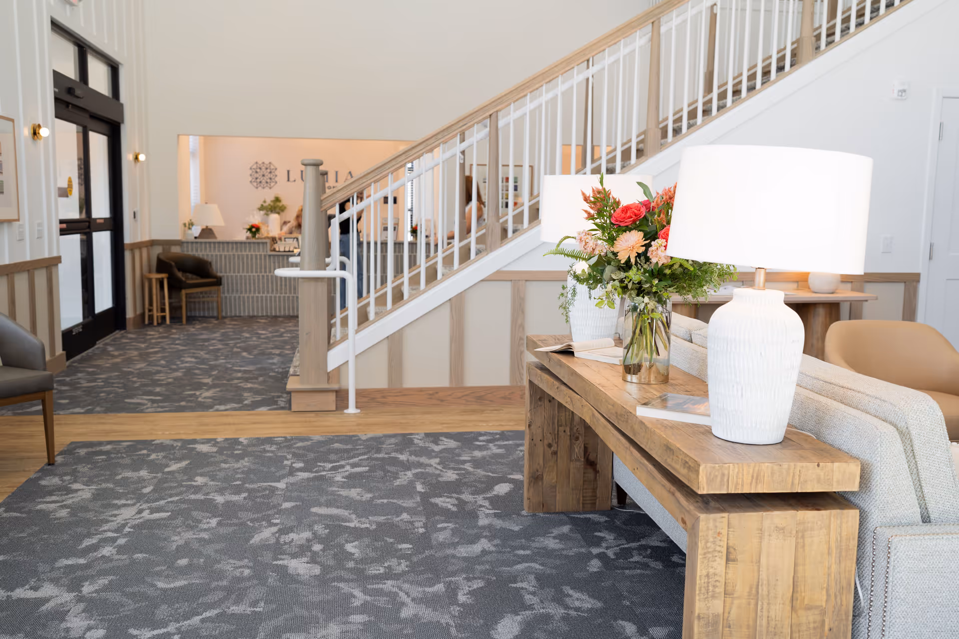 A bright and welcoming interior of an assisted living facility featuring a wooden console table with a white lamp and a vase of colorful flowers. In the background, there is a staircase with white railings, a seating area with chairs, and a reception desk with the Lumia logo on the wall behind it.