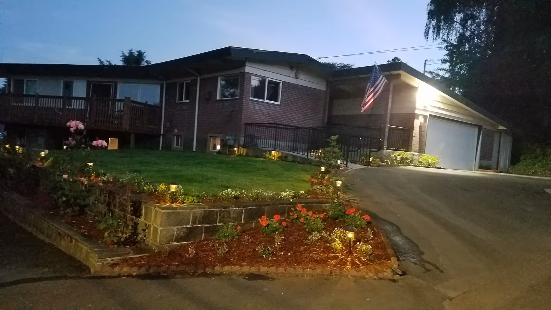 Exterior view of a single-story brick building at dusk with a well-maintained garden featuring flowers and small lights along a stone retaining wall. An American flag is mounted near the entrance, and a driveway leads to a garage with a closed door.