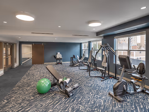 A fitness room with exercise equipment including a recumbent bike, weight machines, and a green exercise ball on a patterned carpet. Large windows on one side let in natural light and show a view of nearby buildings.