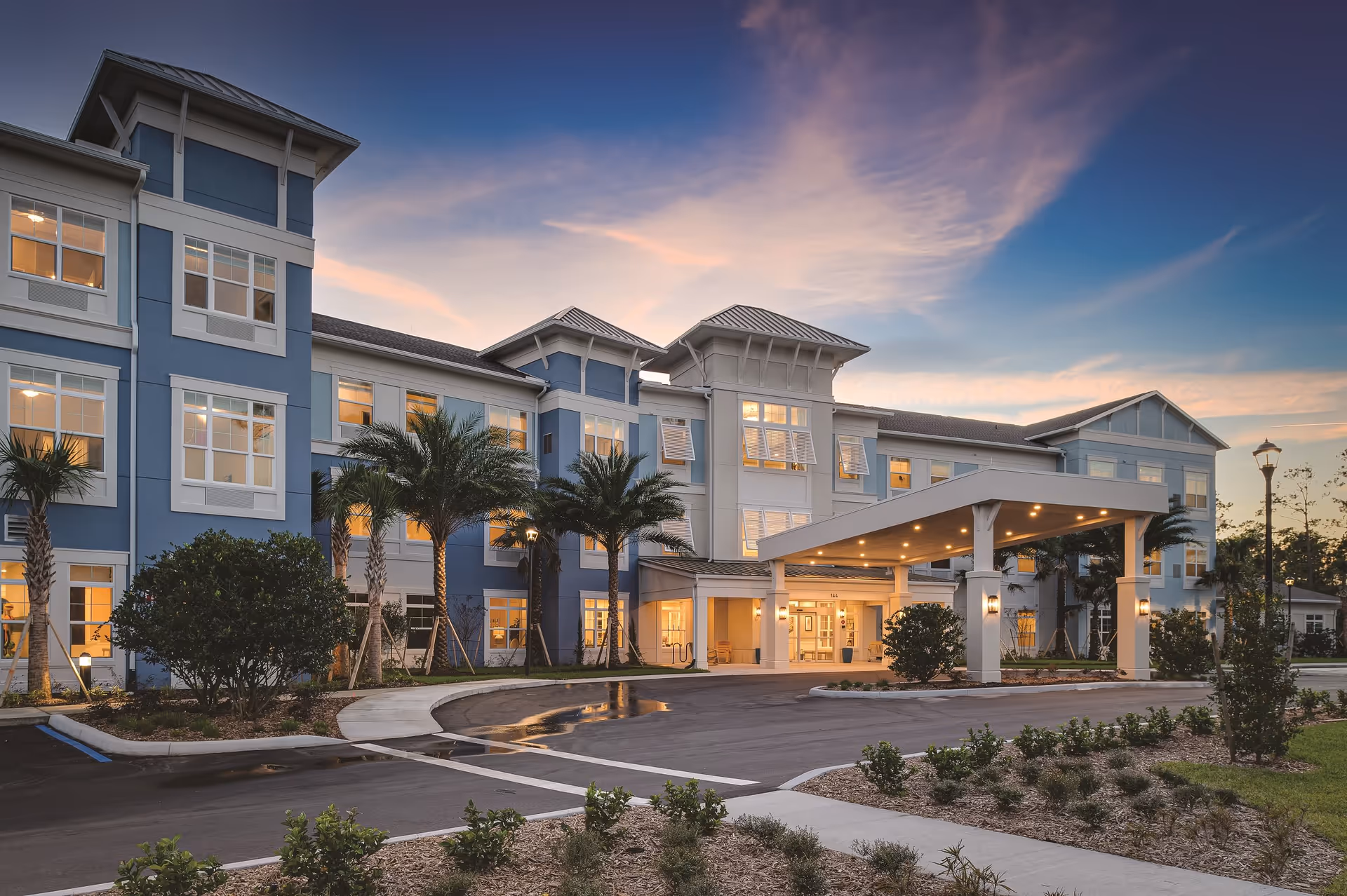 Exterior view of a three-story senior living facility building named Sancerre at Palm Coast during sunset. The building features blue and white walls, multiple windows with lights on inside, a covered entrance with pillars, palm trees, landscaped bushes, and a paved driveway with parking spaces.