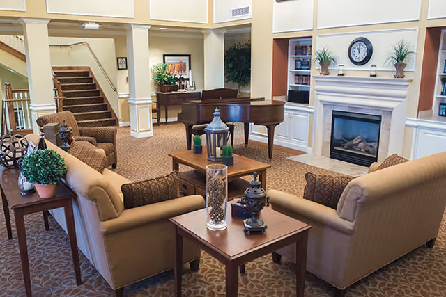 A cozy living room area in a senior living facility featuring two beige sofas facing each other with a wooden coffee table in between. The room includes a fireplace with a clock and plants on the mantel, built-in bookshelves, a grand piano in the background, and a staircase leading upstairs. The space is warmly lit and decorated with plants and framed artwork.